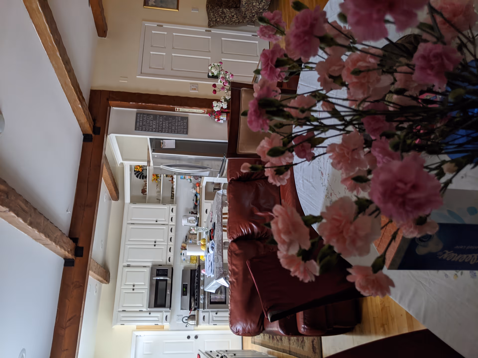 Interior view of a cozy living and kitchen area with wooden ceiling beams. In the foreground, there is a table with a vase of pink flowers and a box of tissues. A red leather sofa and dining chairs are visible, along with a kitchen featuring white cabinets, a stainless steel refrigerator, microwave, and stove. The space is warmly lit and decorated with small plants and wall art.