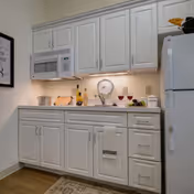 A small kitchen area with white cabinets and drawers, a white microwave mounted above the counter, a white refrigerator on the right, and various kitchen items on the countertop including a pot, cutting board, bottles, a scale, and two glasses of red wine.