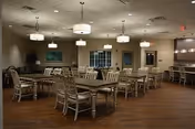 A spacious dining room with multiple wooden tables and chairs arranged neatly. The room features warm lighting from ceiling fixtures, a large window, and neutral-colored walls with a framed artwork. The floor is wooden, and the overall ambiance is calm and inviting.
