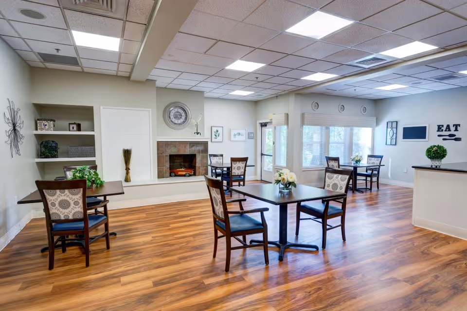 Bright communal dining room with several tables and chairs, wood floors, windows, and a decorative fireplace.