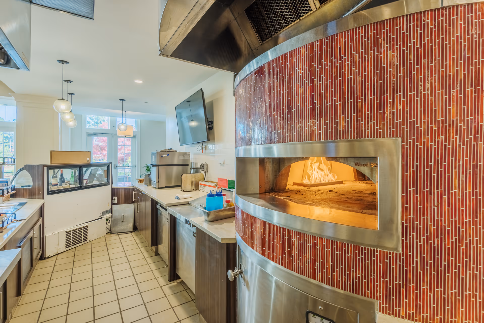 Interior view of a commercial kitchen featuring a large wood-fired oven with a red tiled exterior and stainless steel trim. The kitchen has tiled flooring, countertops with various kitchen equipment, a mounted TV, and large windows letting in natural light.