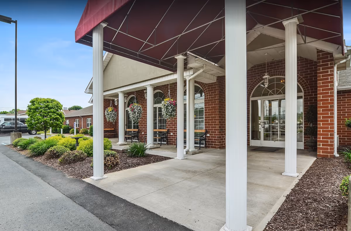 Entrance of a brick building with white columns supporting a maroon awning. There are hanging flower baskets and benches along the walkway leading to glass double doors. Landscaping with green shrubs and a small tree is visible along the side.