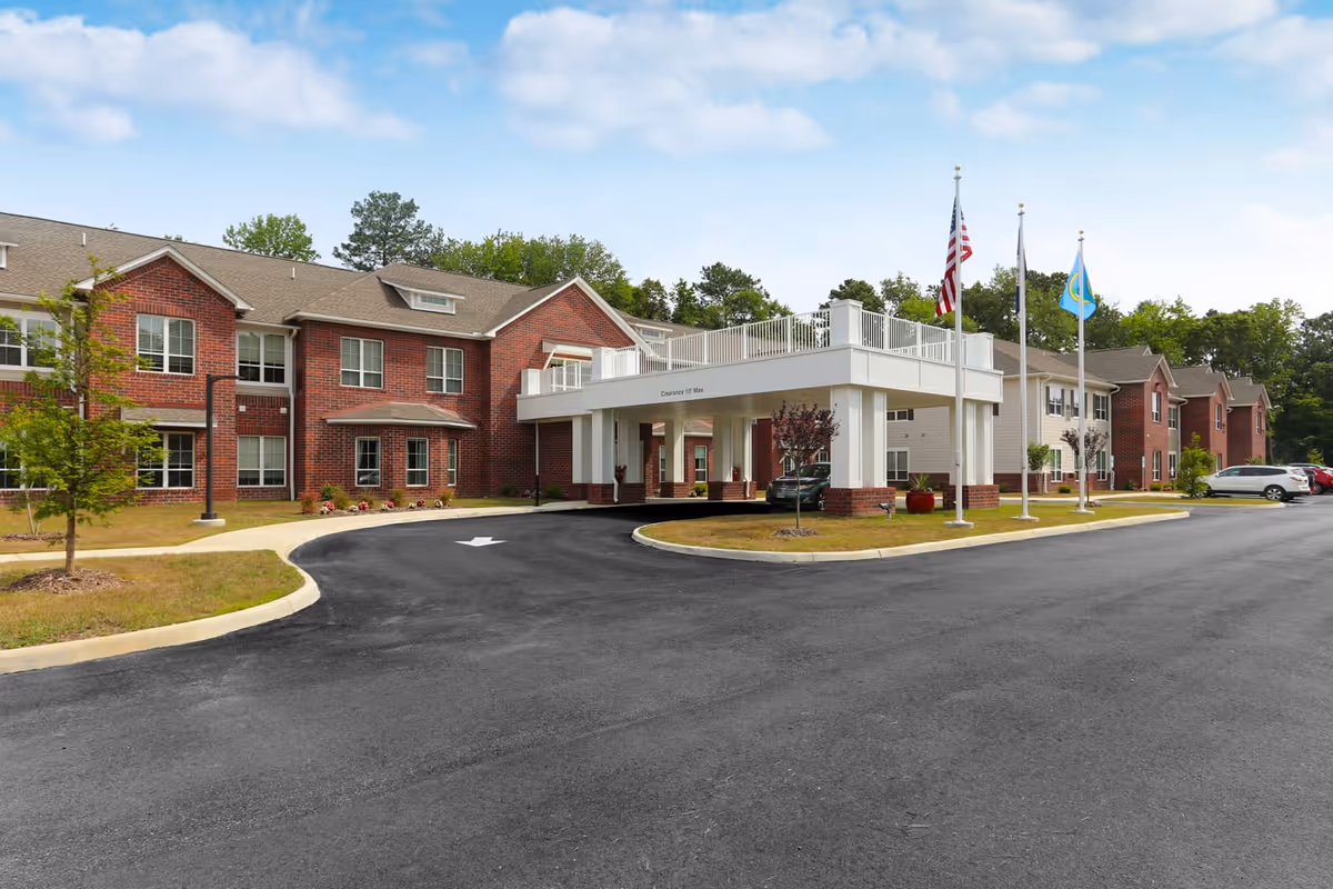 Exterior view of a two-story senior living facility building with red brick and beige siding. The building has multiple windows and a covered entrance with white pillars. There are three flagpoles with flags near the entrance, a paved driveway with a white arrow, and some parked cars. Trees and greenery surround the building under a partly cloudy sky.
