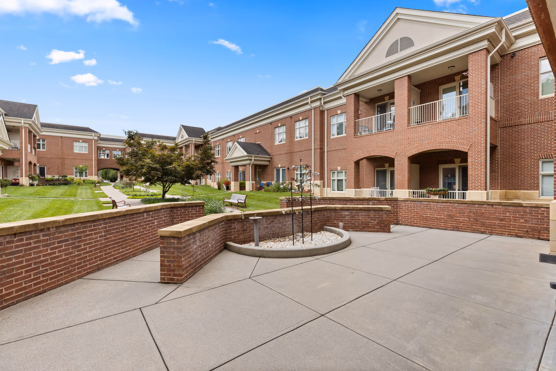 Outdoor courtyard area of a senior living facility with brick buildings surrounding a paved patio and green lawn with benches and a tree under a blue sky with some clouds.
