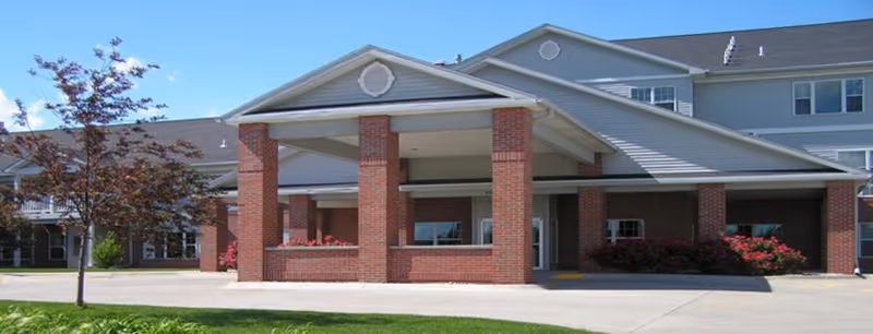 Front exterior view of Kinship Pointe Northridge facility showing a covered entrance with brick pillars, a driveway, manicured lawn, and a multi-story building with multiple windows under a clear blue sky.