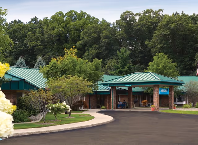 Front exterior of Valley Pines Senior Living with green roof, brick columns, and surrounding trees.