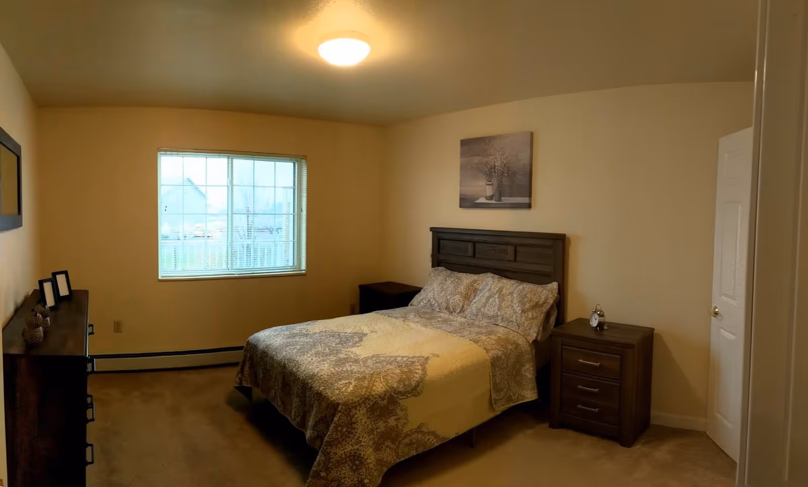 A bedroom with a double bed covered in a patterned quilt, two matching nightstands on either side of the bed, a dresser with decorative items, a window with blinds, beige walls, and a ceiling light fixture.