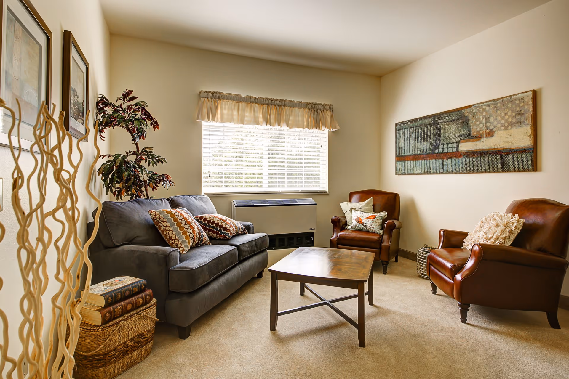A cozy living room with a gray sofa adorned with patterned pillows, two brown leather armchairs with decorative cushions, a wooden coffee table in the center, a large window with blinds and a valance, a tall plant in the corner, and framed artwork on the walls.