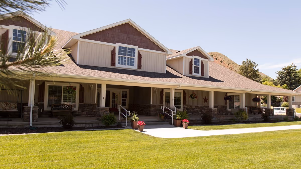 Exterior view of a single-story assisted living facility building with a covered porch, potted plants, benches, and a well-maintained lawn in front. The building has beige siding with stone accents and multiple windows with shutters. Trees and hills are visible in the background under a clear sky.