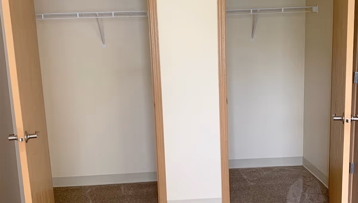 Two empty closets with open wooden doors, each closet has a white hanging rod and beige walls with carpeted floors.