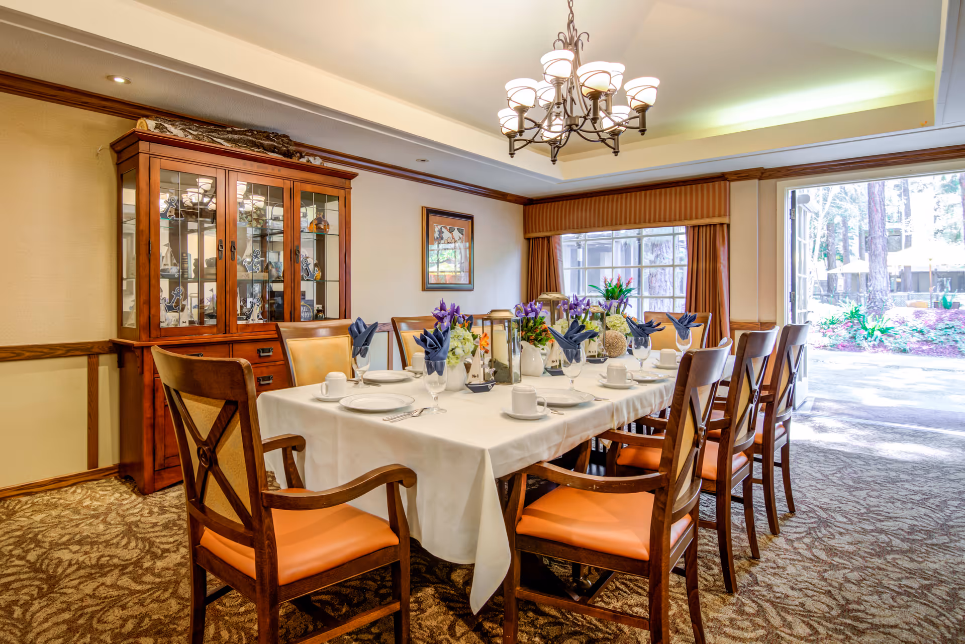 A well-lit dining room with a long table set for a meal, featuring white tablecloth, plates, cups, glasses, and blue folded napkins. The room has wooden chairs with orange cushions, a wooden cabinet with glass doors displaying decorative items, a chandelier hanging from the ceiling, and large windows with brown curtains letting in natural light and showing an outdoor garden area.