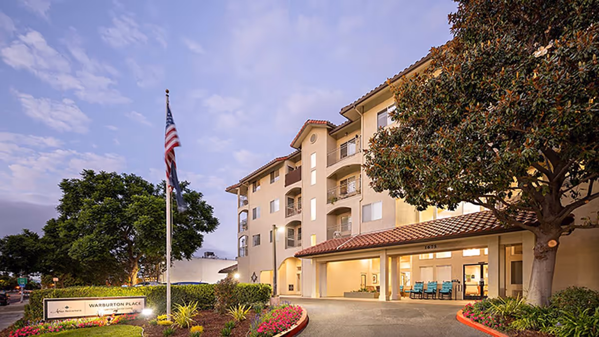 Exterior view of Holiday Warburton Place senior living facility at dusk, showing a multi-story building with balconies, a covered entrance, landscaped garden with flowers and trees, and an American flag on a flagpole.