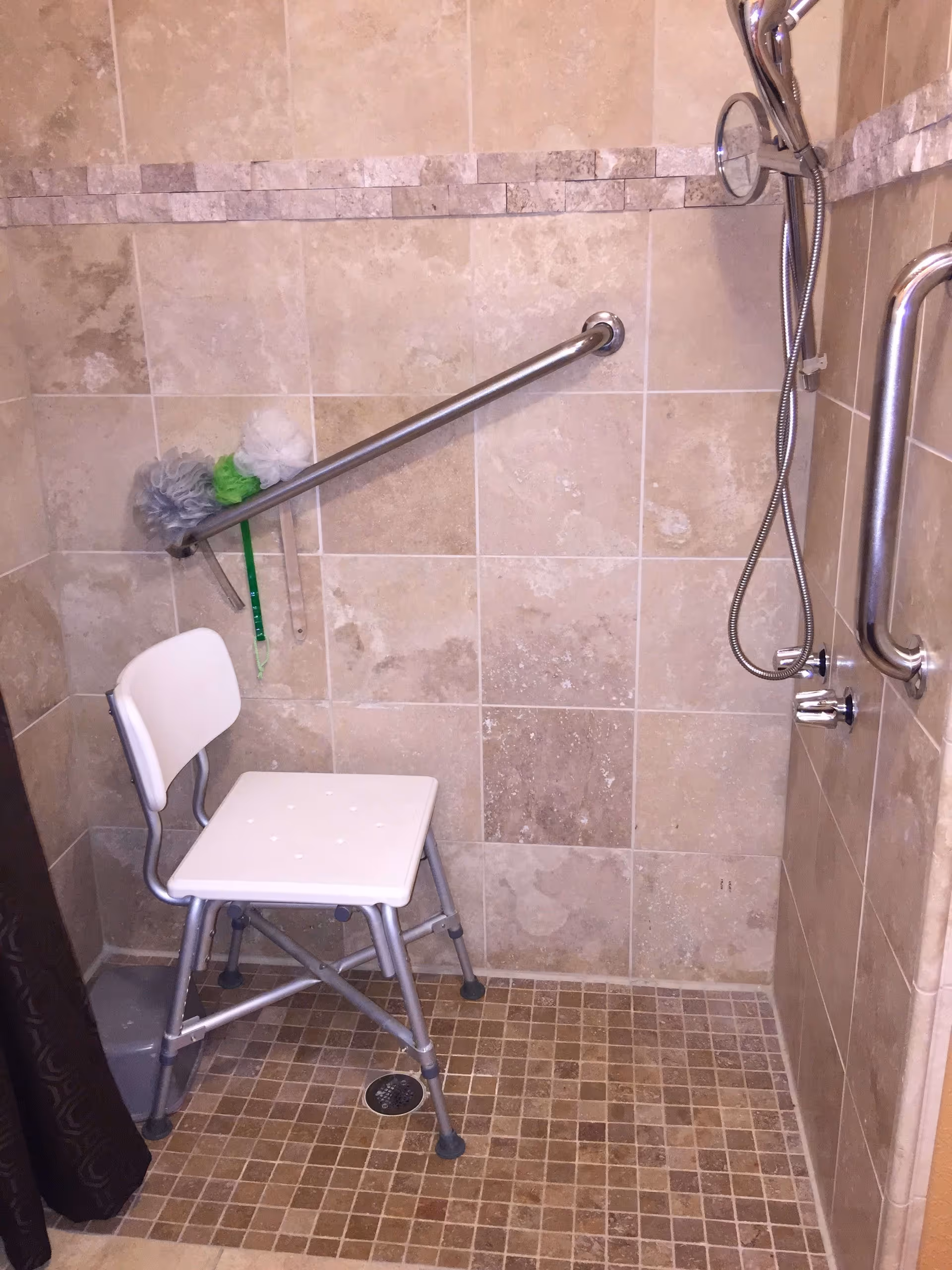 A tiled shower area with beige and brown tiles, featuring a white shower chair with metal legs, a handheld showerhead attached to the wall, and two metal grab bars for support. There are three bath poufs hanging on the grab bar on the left side.