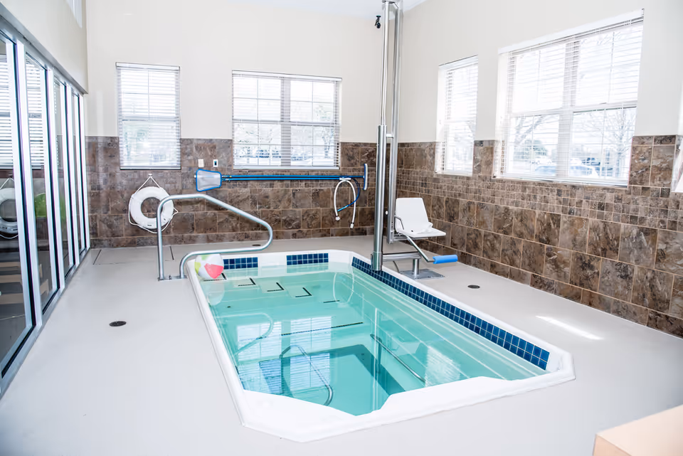Indoor therapy pool with clear water, surrounded by tiled walls and windows letting in natural light. The pool has a metal handrail, a life preserver hanging on the wall, a pool cleaning net, and a white pool lift chair for accessibility.