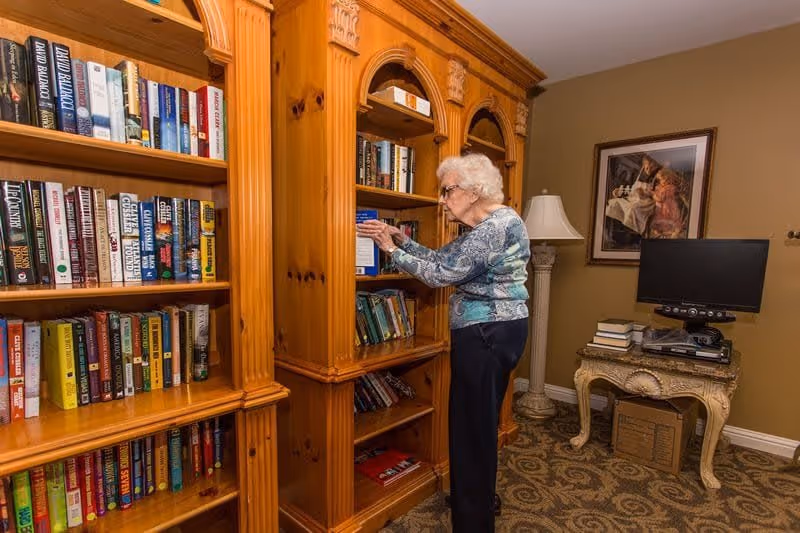 An elderly woman with white hair and glasses is standing and reaching for a book on a wooden bookshelf filled with various books in a cozy room. The room has a patterned carpet, a small ornate table with a stack of books and a TV on it, a floor lamp, and a framed painting on the wall.