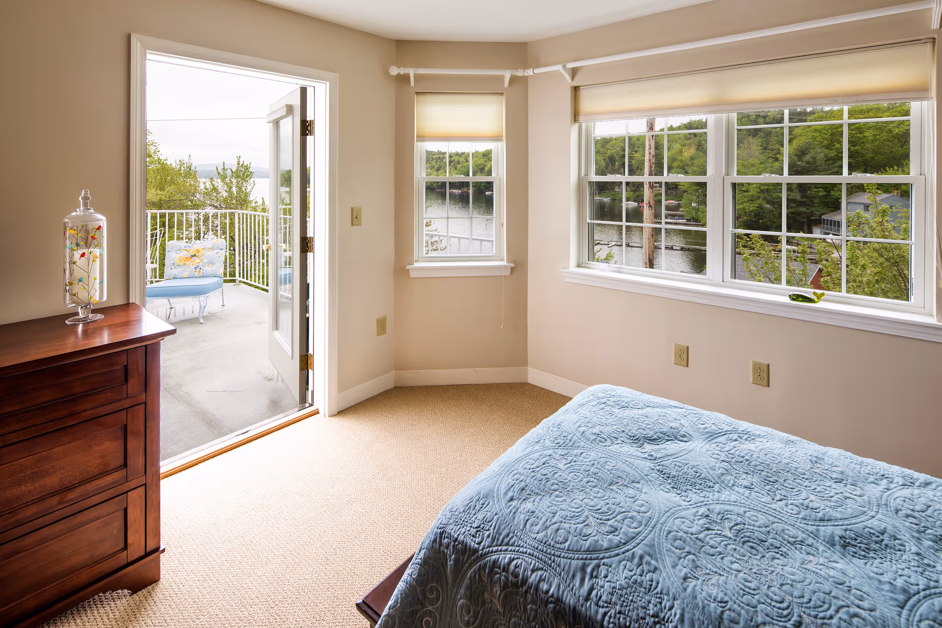 Bedroom with a blue quilted bed, wooden dresser and open door to a balcony overlooking a lake.