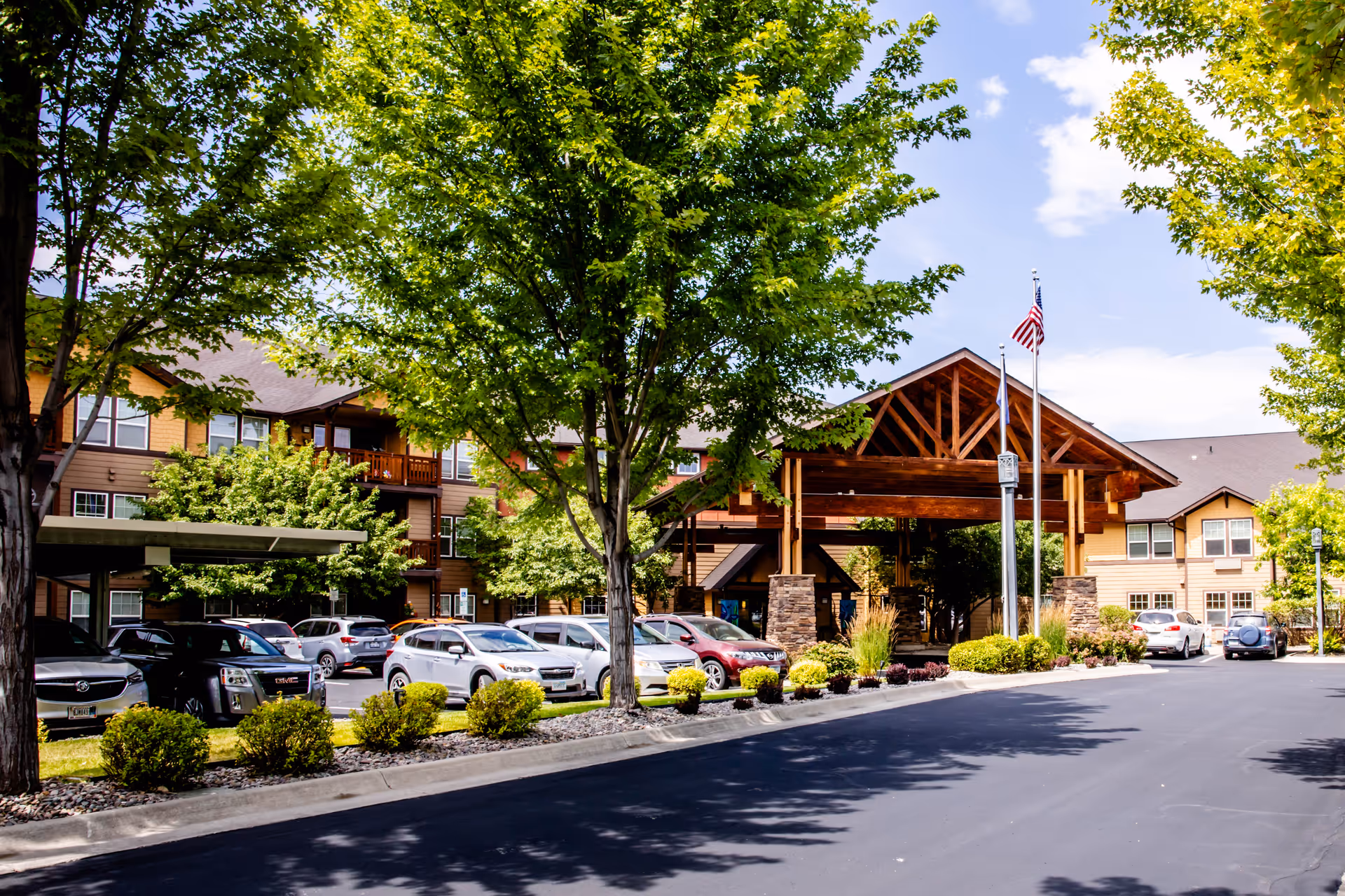 Front entrance and parking area of a senior living facility with a covered wooden porte-cochère, flagpole, parked cars, and trees.