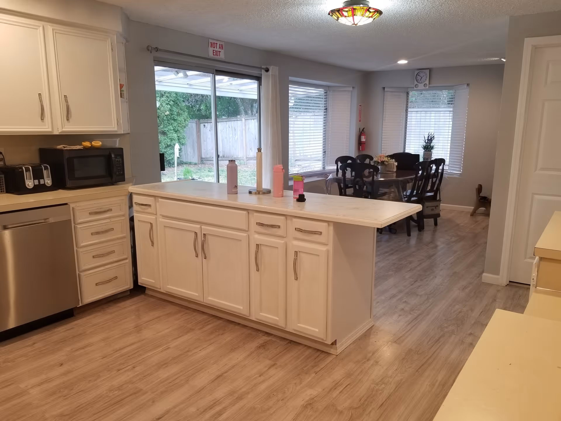 Interior view of a kitchen and dining area in a senior living facility. The kitchen features white cabinets, a dishwasher, a microwave, a toaster, and a countertop island with water bottles and a paper towel holder. The dining area has a round table with six chairs, a flower centerpiece, and large windows with blinds. A sliding glass door leads to an outdoor space with a wooden fence and greenery.