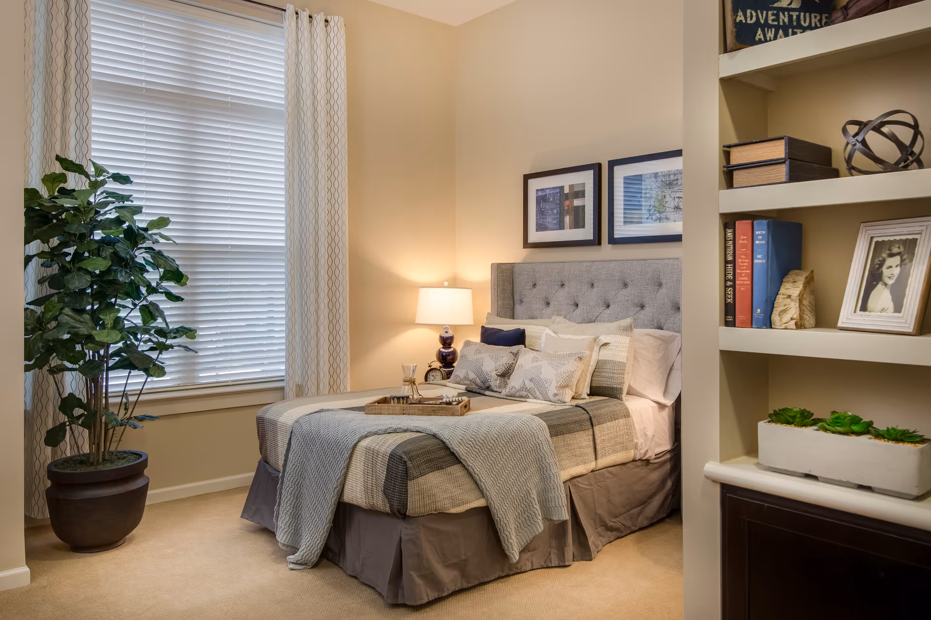 A cozy bedroom with a neatly made bed featuring multiple pillows and a gray quilt. A bedside table with a lamp is next to the bed. There is a large window with white blinds and patterned curtains. A tall potted plant is placed near the window. On the right side, built-in shelves hold books, decorative items, a framed photo, and a planter with succulents.