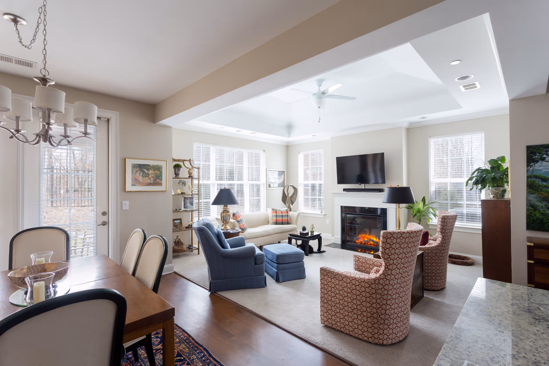 A bright and spacious living room with large windows allowing natural light to fill the space. The room features a beige sofa with colorful pillows, two patterned armchairs, and a blue armchair with an ottoman. A fireplace with a mounted flat-screen TV above it is centered on one wall. There are two black table lamps on side tables, a ceiling fan, and various plants and decorative items around the room. Adjacent to the living area is a dining table with chairs and a chandelier overhead.