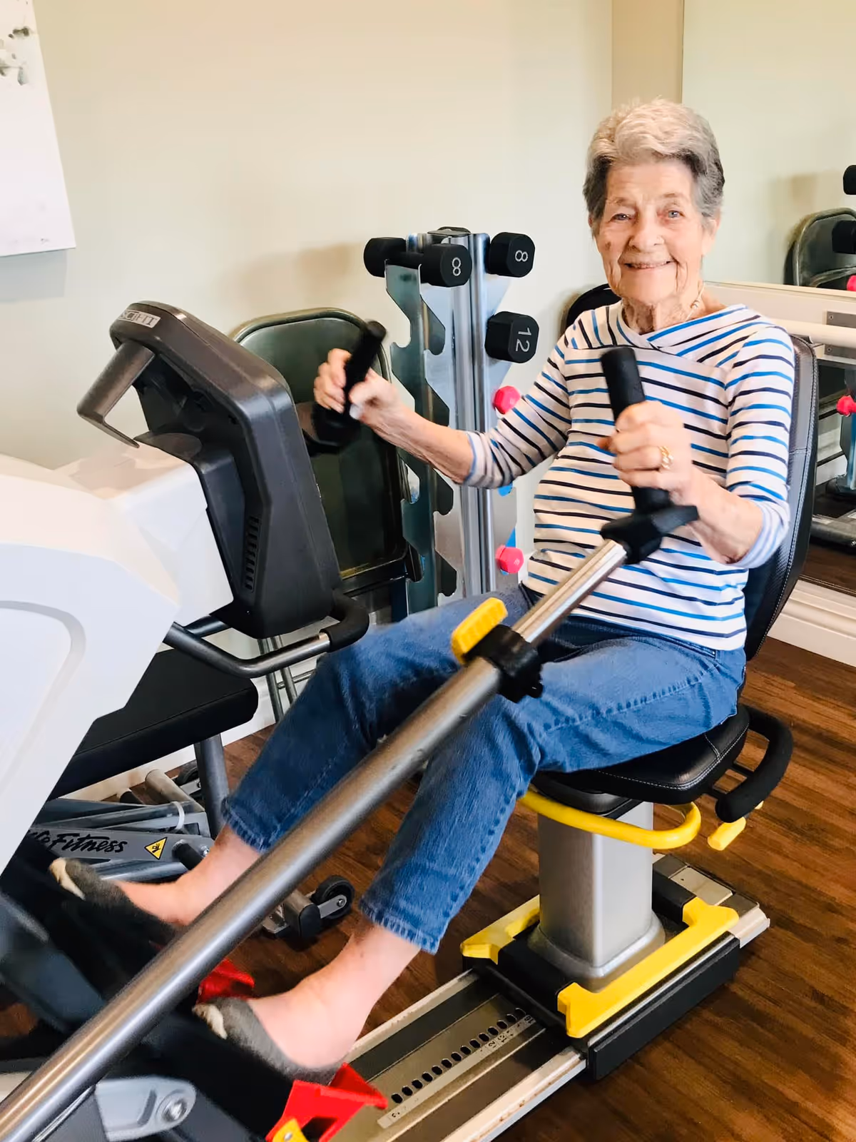 An elderly woman wearing a striped shirt and blue jeans is seated on a rowing exercise machine in a fitness room. She is smiling and holding the handles of the machine, with weights visible in the background.