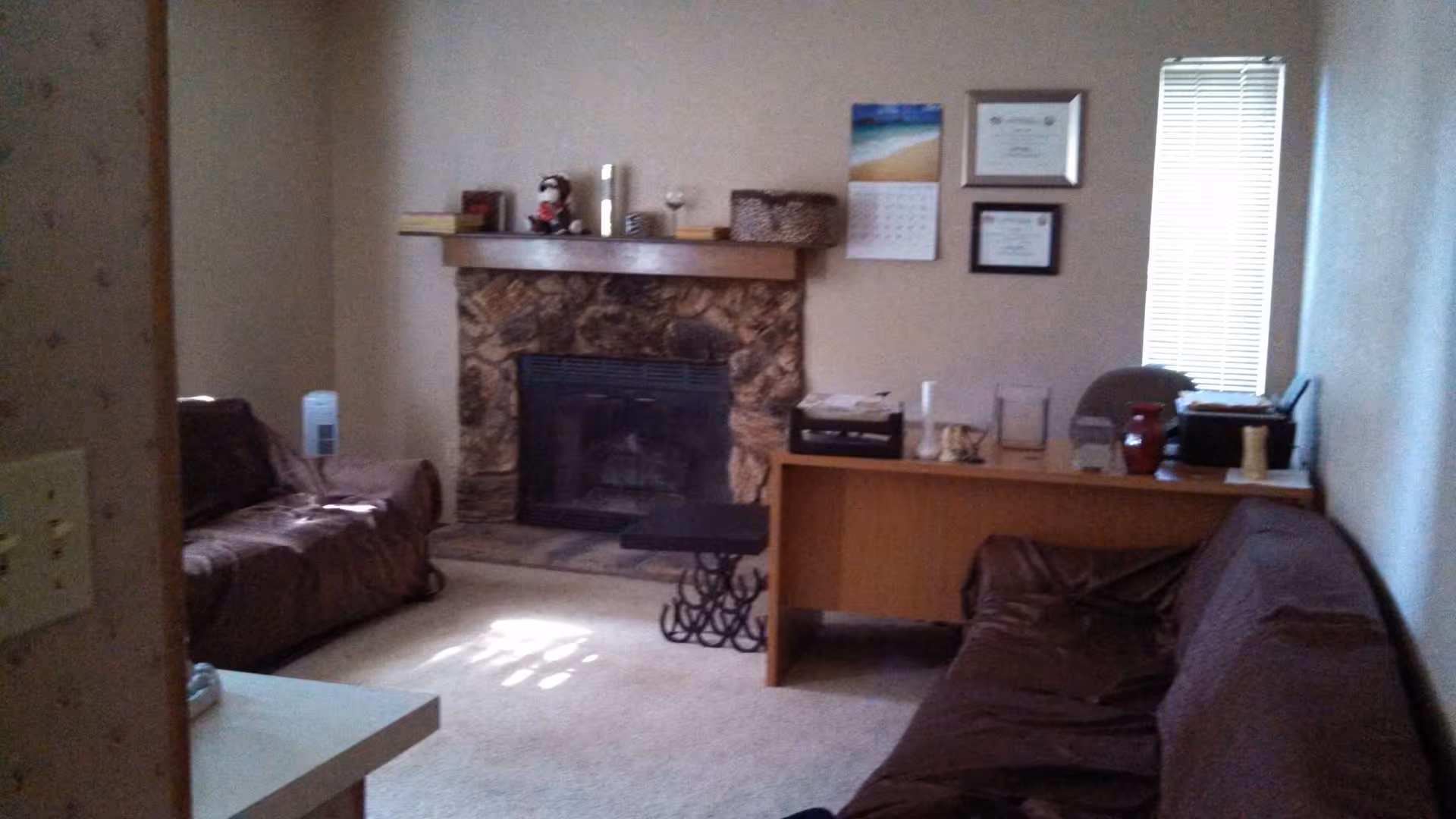 A small living room with two brown couches facing each other, a stone fireplace with a wooden mantle decorated with small items, a wooden desk with various objects on it, and a window with blinds letting in light.