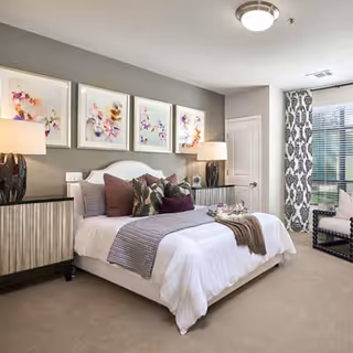 A well-decorated bedroom featuring a large bed with white bedding, multiple pillows, and a throw blanket. On either side of the bed are matching nightstands with lamps. Above the bed are four framed abstract floral artworks. To the right, there is a window with patterned curtains and a cushioned chair beside it. The room has a neutral color palette with beige carpet and light gray walls.