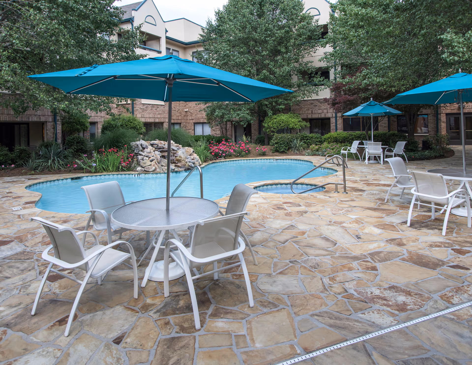 Outdoor pool area at Delmar Gardens of Gwinnett Senior Living featuring a kidney-shaped pool with a small rock waterfall, surrounded by stone tile flooring. Several white chairs and tables with blue umbrellas are arranged around the pool. The background shows a brick building with windows and lush green trees and shrubs.