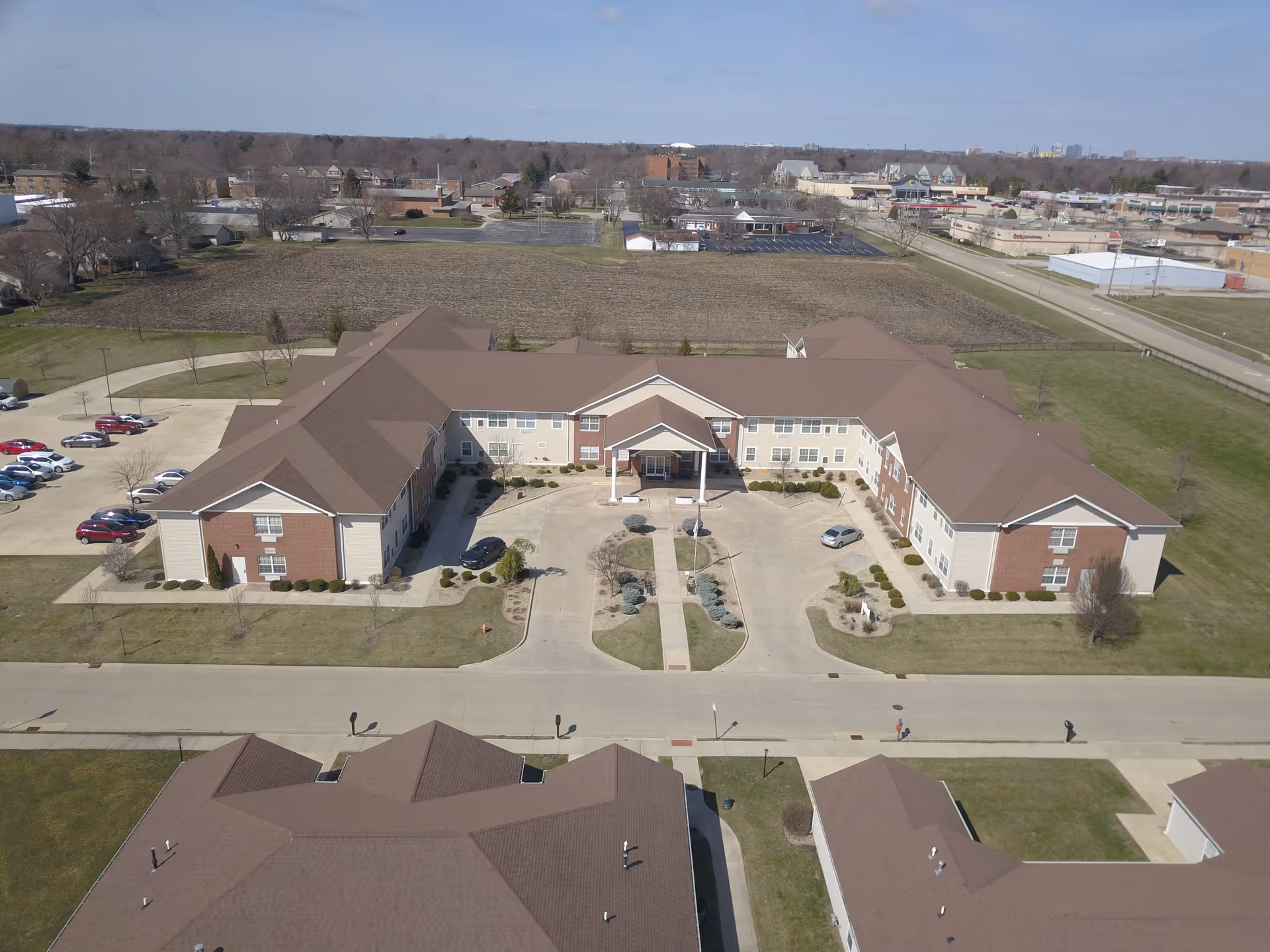 Aerial view of Prairie Winds of Urbana senior living facility showing a large, U-shaped building with a brown roof and beige walls with brick accents. The building is surrounded by well-maintained lawns, parking lots with several cars, and a paved driveway leading to the main entrance with a covered portico. In the background, there are fields, trees, and other buildings under a clear blue sky.