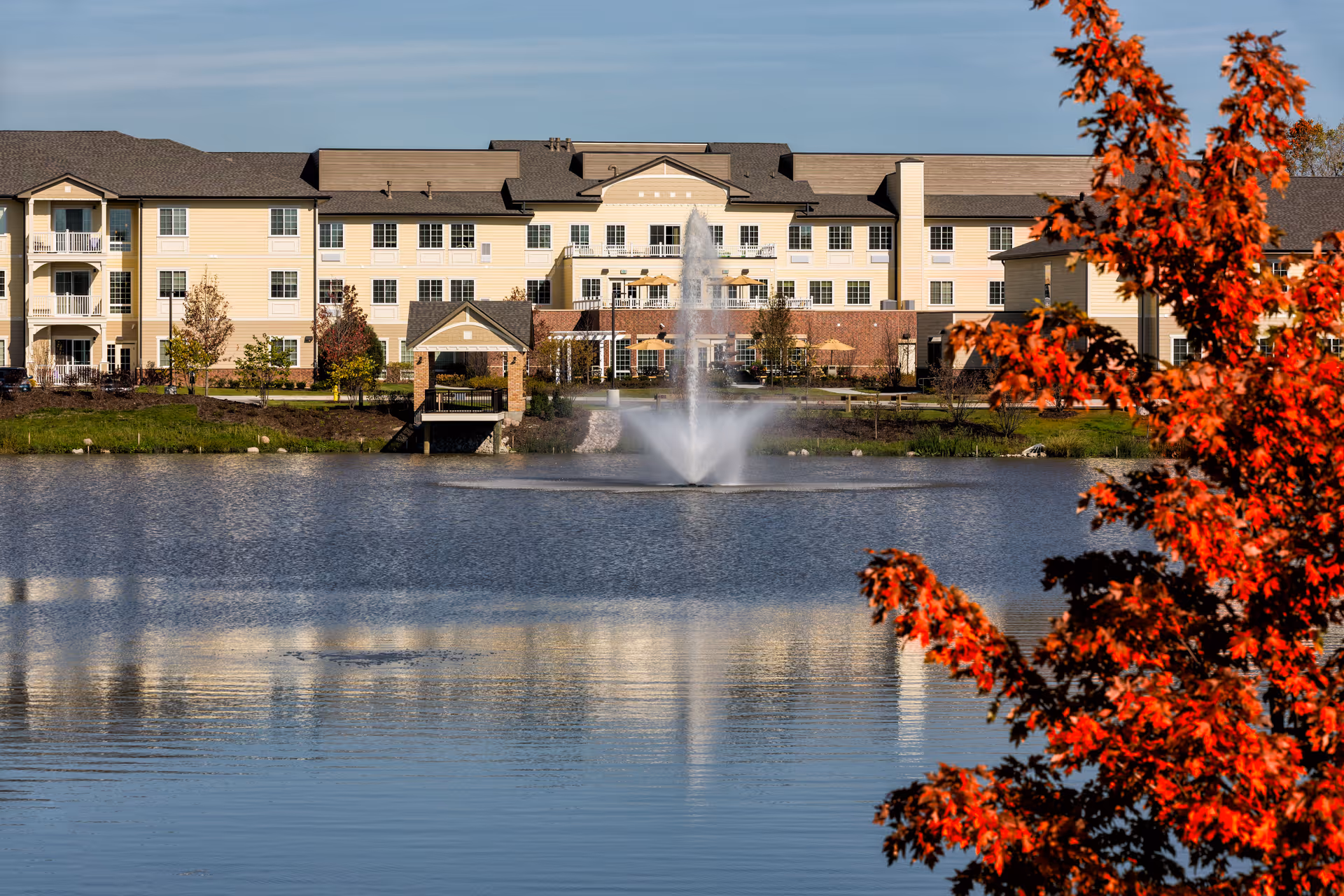 A large senior living facility building with multiple windows and balconies is visible across a pond with a water fountain in the center. The foreground features a tree with bright red autumn leaves, and the sky is clear and blue.