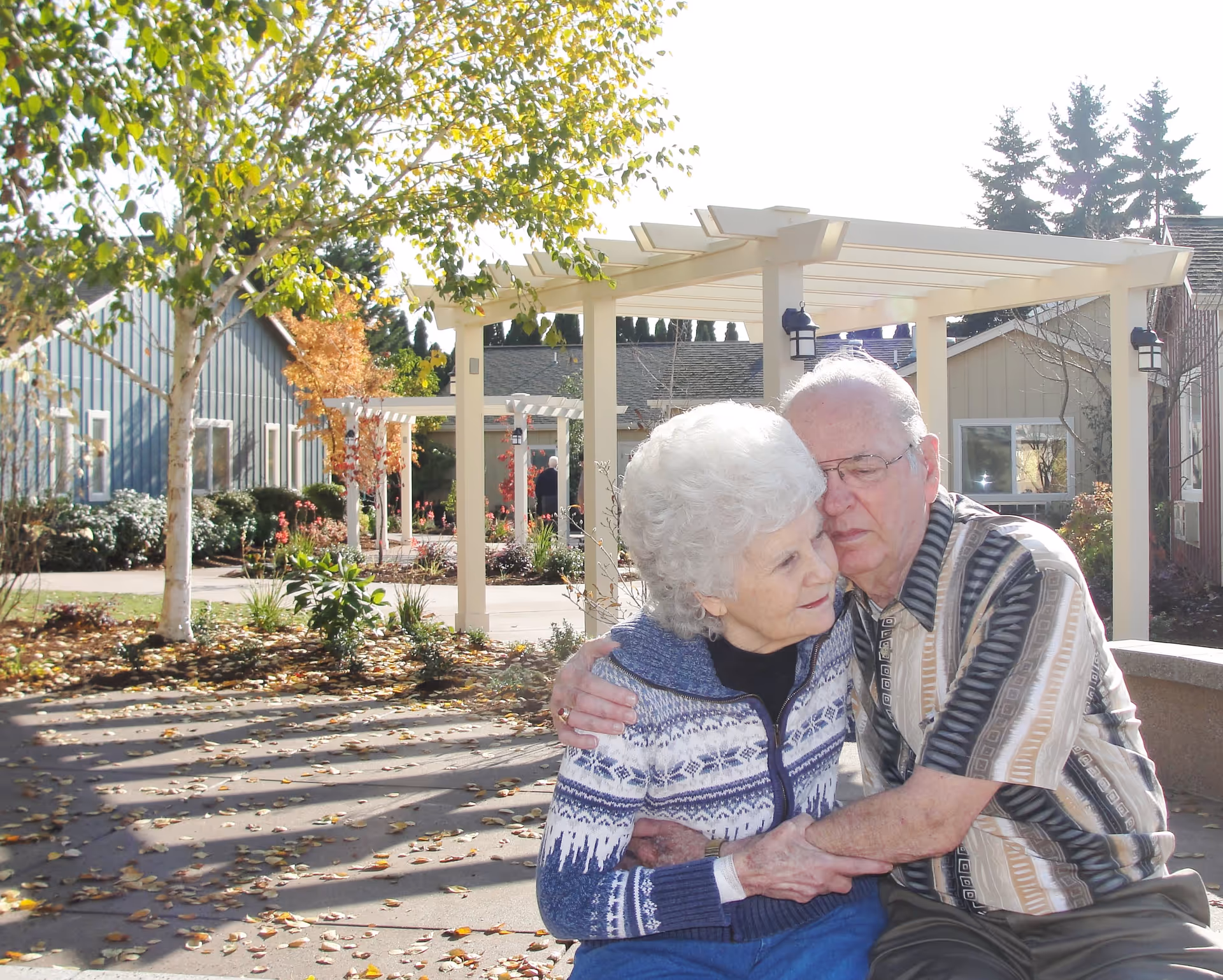 An elderly couple sitting on a bench outdoors in a garden area of a senior living facility. The man is hugging the woman affectionately. The background shows trees, plants, and pergolas with buildings behind them.