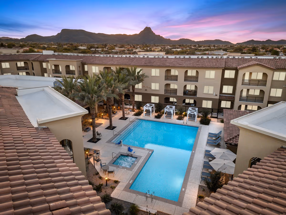Aerial view of a senior living courtyard with a central swimming pool, hot tub, palm trees, and surrounding multi-story building at sunset with mountains in the background.