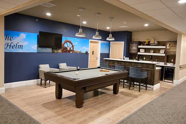 A recreational room with a pool table in the center, two armchairs against a navy blue wall with a nautical-themed mural and a mounted TV. To the right, there is a bar area with three bar stools, shelves with decorative items, and a small kitchenette.