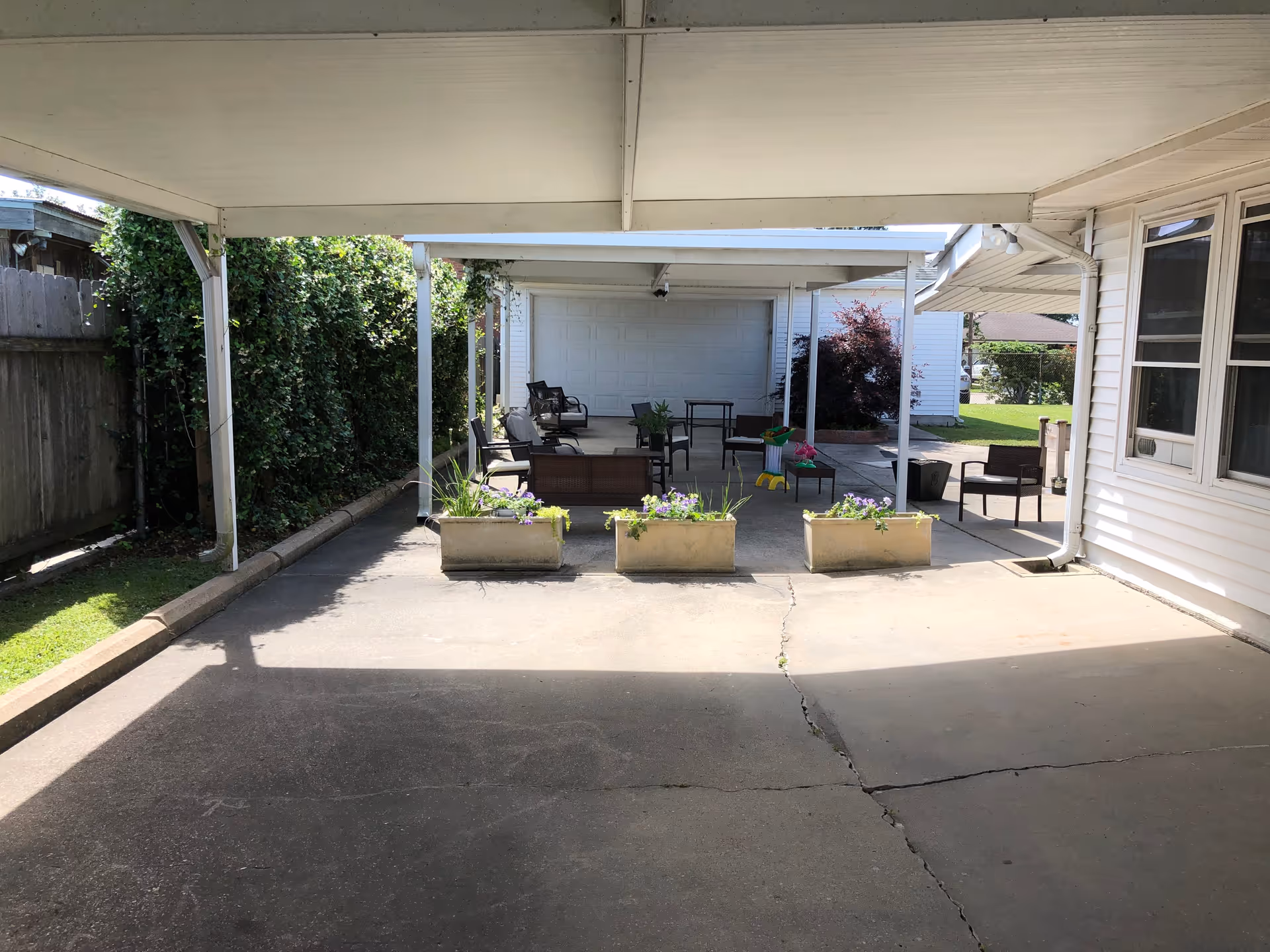 Covered outdoor patio area with several chairs and tables arranged for seating. Three rectangular planters with flowers are placed in front of the seating area. The patio is adjacent to a white building with windows and a garage door in the background. Green bushes and a wooden fence line one side of the patio.