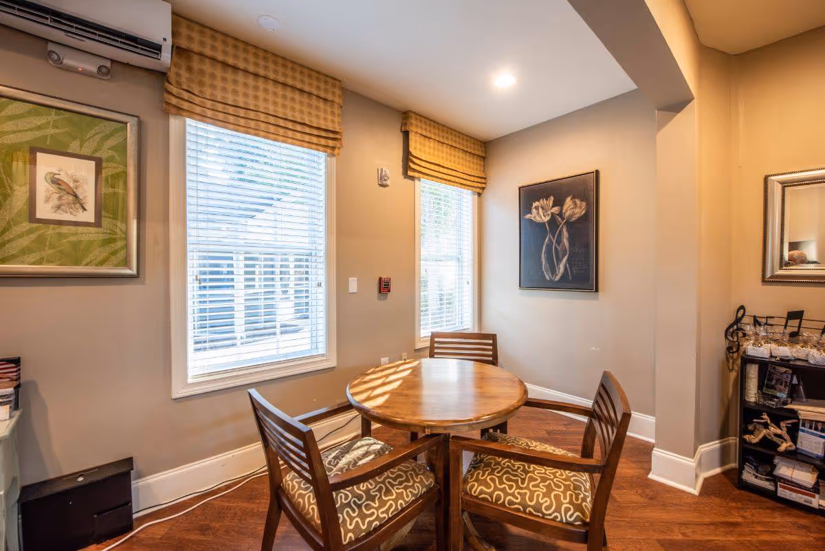 A cozy interior corner of a senior living facility with a round wooden table surrounded by four wooden chairs with patterned cushions. Two windows with blinds and yellow valances let in natural light. The walls are decorated with framed artwork, including a bird and floral prints. A small shelving unit with decorative items and a mirror is visible on the right side.