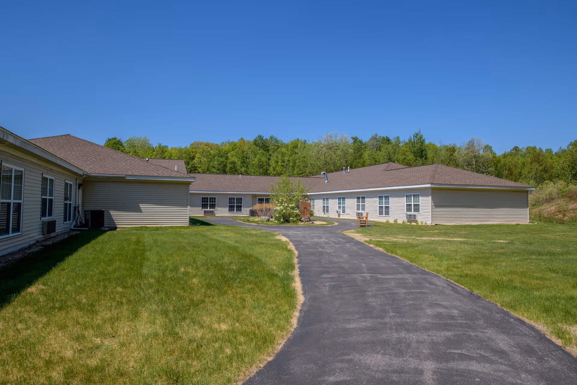 A paved pathway leads through a grassy courtyard area surrounded by single-story beige buildings with multiple windows under a clear blue sky. Trees and greenery are visible in the background.