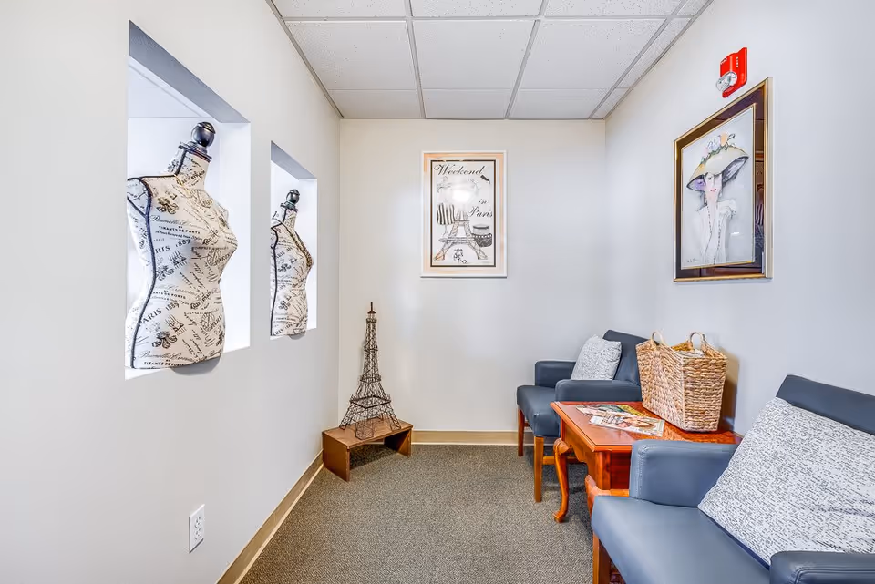 A small waiting or sitting area with two blue armchairs with patterned cushions, a wooden side table with a wicker basket and magazines, and two decorative dress forms mounted in wall niches. The walls are light-colored with framed artwork, including a picture of the Eiffel Tower and a fashion illustration. The floor is carpeted.