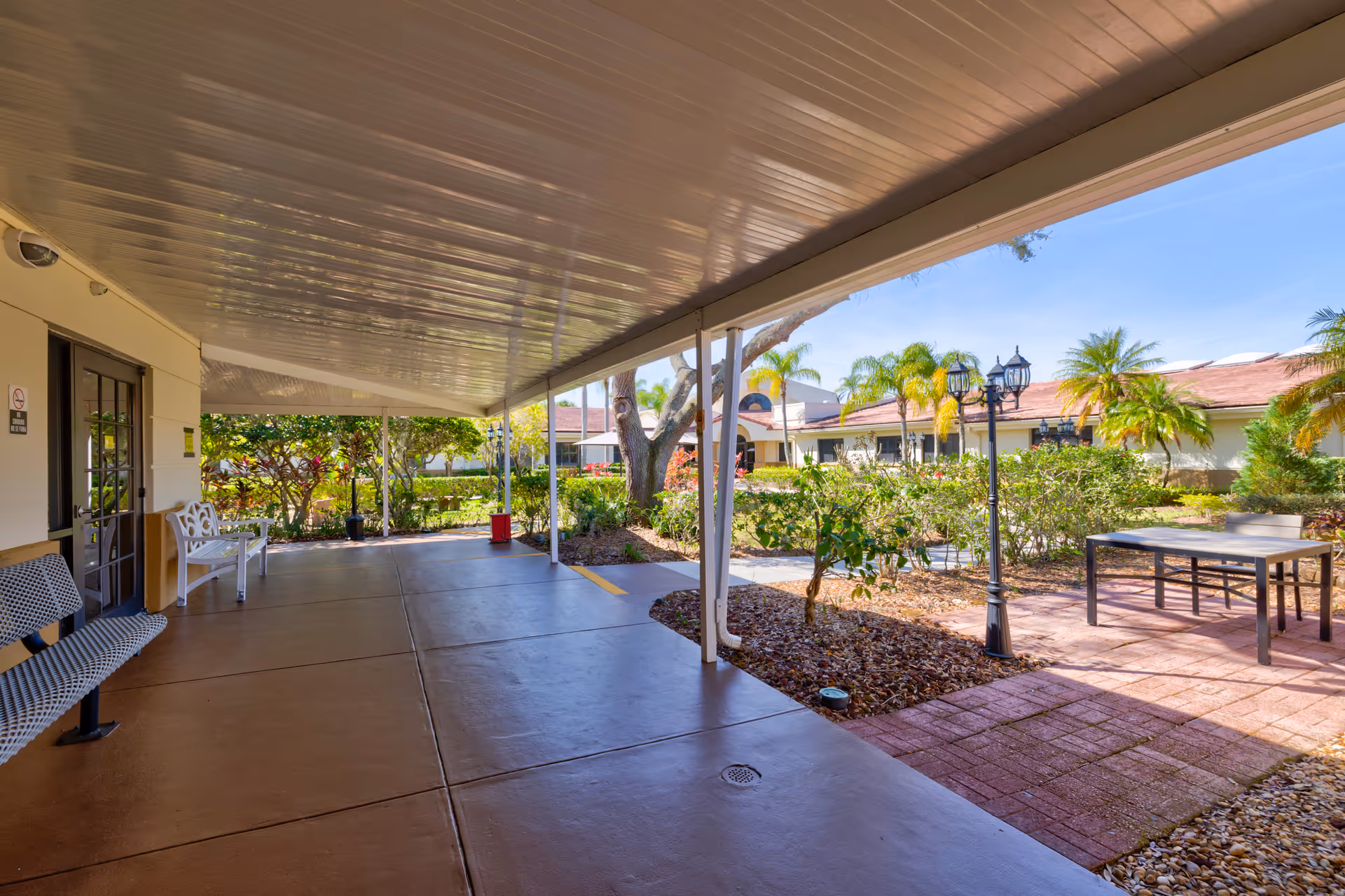 Covered outdoor walkway with benches on the left side, leading to a garden area with trees, shrubs, and a patio with a table and chairs under a clear blue sky.