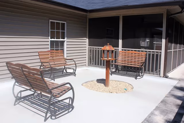 Outdoor patio area with three wooden benches arranged around a central bird feeder on a concrete surface, adjacent to a building with beige siding and screened porch.