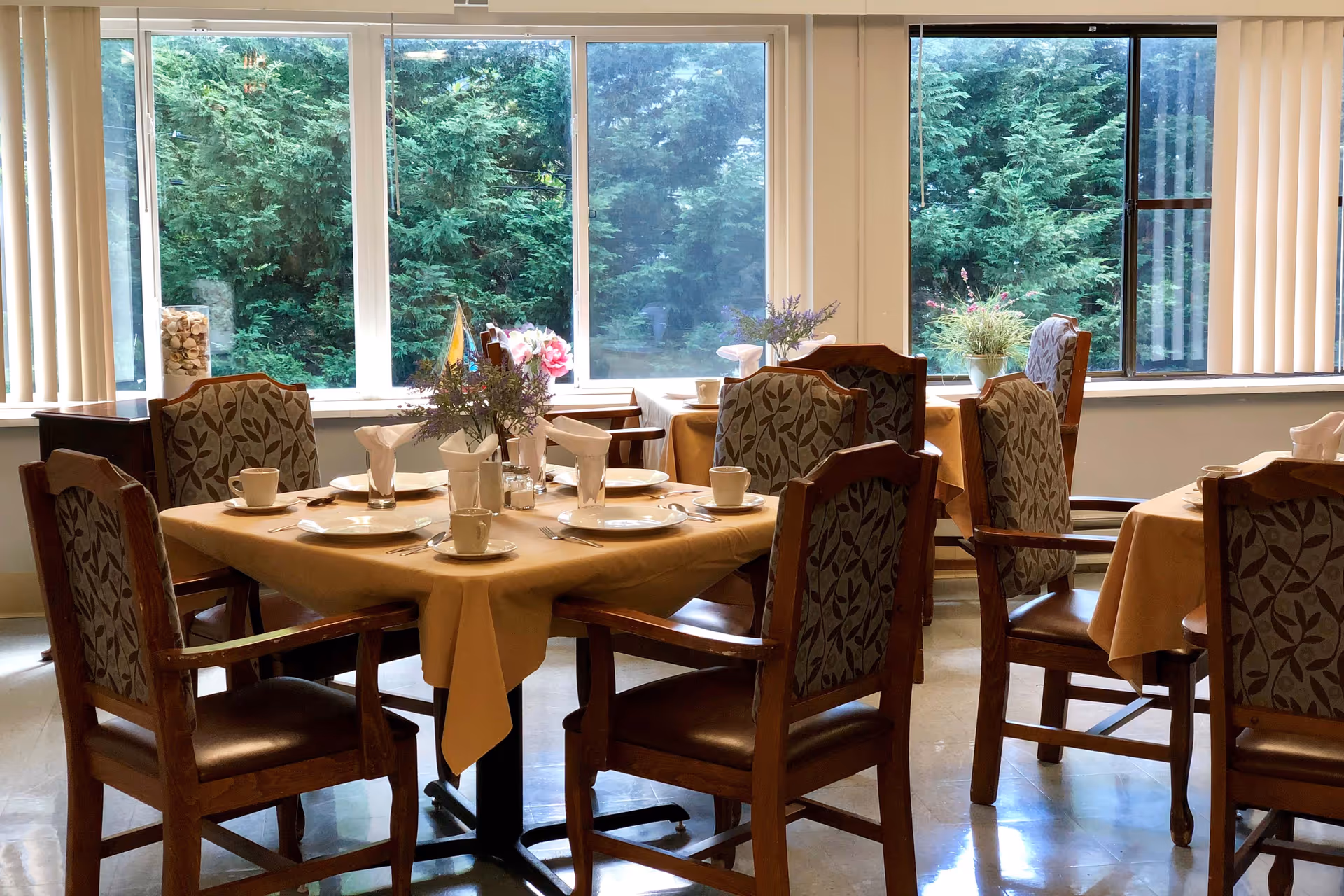 Sunlit dining room with several set tables and wooden chairs facing large windows overlooking greenery.