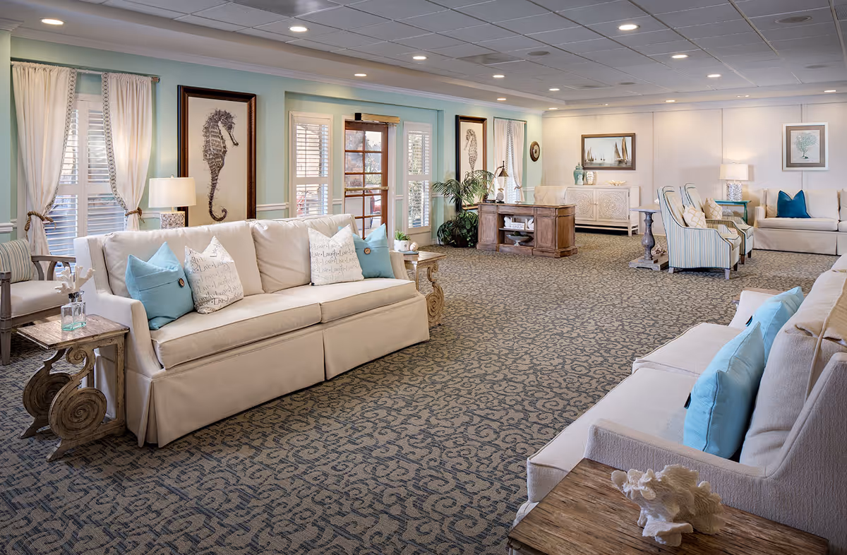 A spacious and well-lit senior living facility common area with beige sofas adorned with blue and patterned pillows, wooden side tables, framed seahorse artwork on the walls, a patterned carpet, and large windows with white curtains allowing natural light to fill the room.