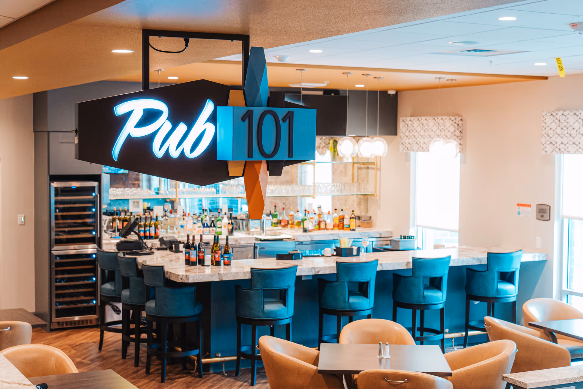 Interior view of a modern pub area named Pub 101 with a marble countertop bar surrounded by teal bar stools. Behind the bar are shelves stocked with various bottles of alcohol and glassware. The space is well-lit with large windows and pendant lights, and there are tables and chairs in the foreground.