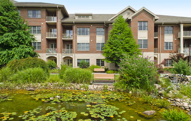 Exterior view of a multi-story brick residential building with balconies, surrounded by lush greenery and trees. In the foreground, there is a pond filled with lily pads and rocks along the edge, with benches placed near the water.
