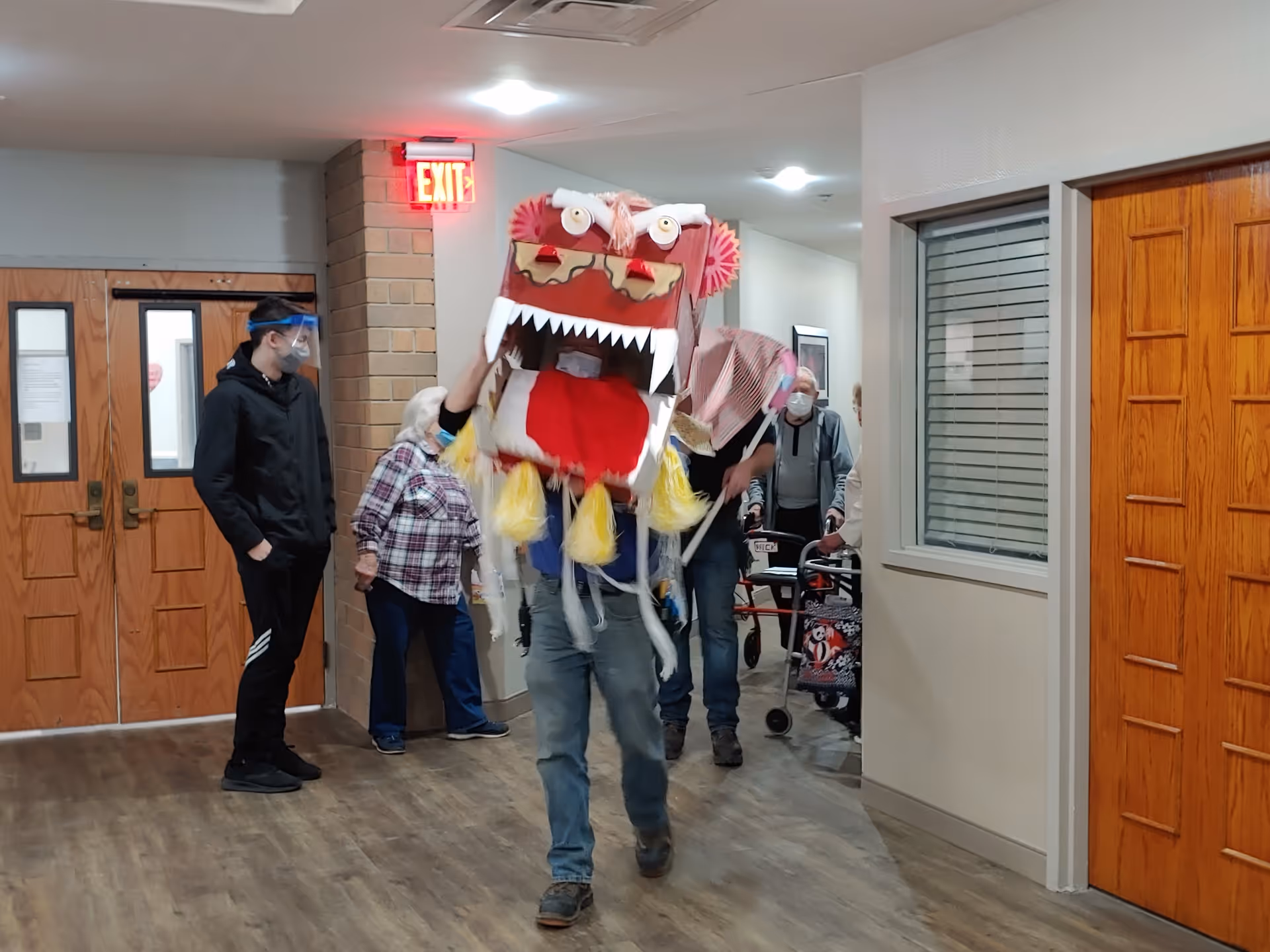 A person wearing a colorful dragon costume walks down a hallway in a supportive living facility. Several elderly people and a younger person wearing a face shield stand nearby, watching the performance. The hallway has wooden doors, a brick column, and an illuminated exit sign.