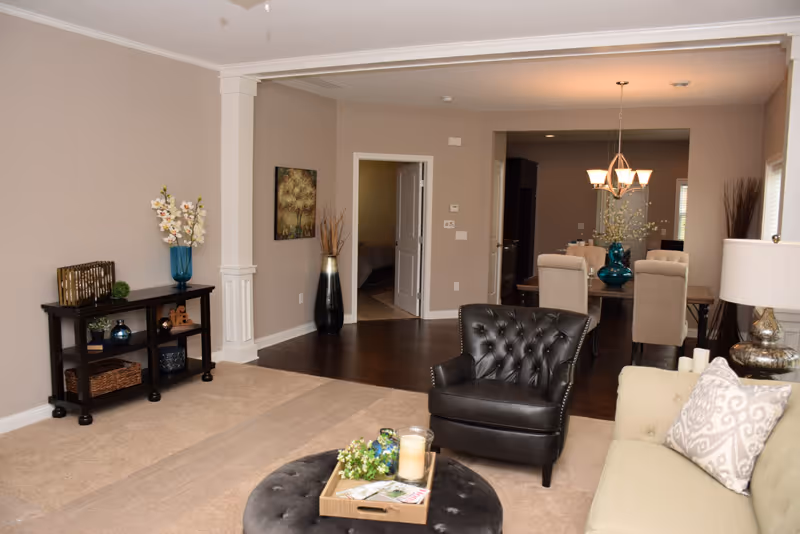 A cozy living room area with a beige sofa adorned with patterned pillows, a black leather armchair, and a round ottoman with a tray holding a candle and some greenery. The room has beige walls and carpeted flooring, with a dark wooden console table against one wall decorated with a blue vase of flowers and other decorative items. In the background, there is a dining area with a wooden table and beige upholstered chairs under a chandelier, and an open doorway leading to another room.