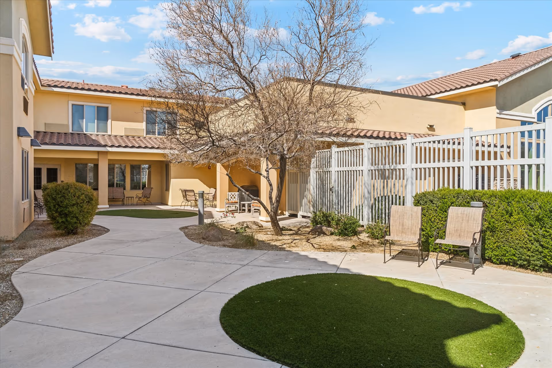 Outdoor courtyard area at The Groves Assisted Living & Memory Care featuring a paved walkway, a leafless tree, green bushes, two chairs, and a beige building with a tiled roof under a blue sky with some clouds.