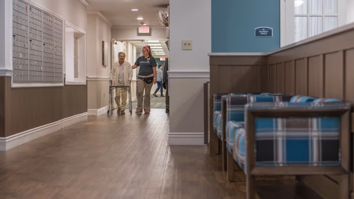 A hallway in a senior living facility with wooden flooring and mailboxes on the left wall. An elderly woman using a walker is being assisted by a staff member wearing a Kingston Residence shirt. On the right side, there are blue and plaid cushioned chairs along a wooden partition. A sign on the wall reads 'Business Office'.