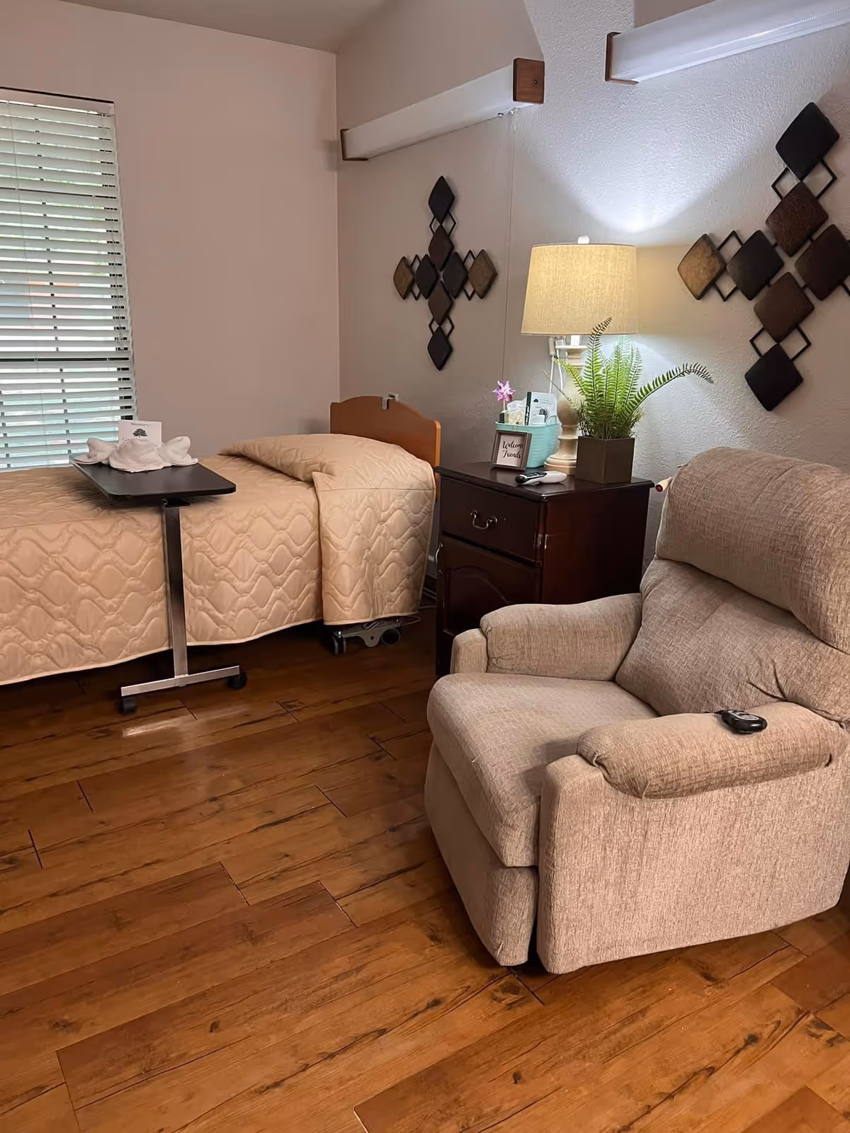 A cozy room with a single bed covered in a beige quilt, a small overbed table with folded towels, a wooden nightstand with a lamp, a small plant, and decorative items. There is a comfortable beige recliner chair in the foreground and wall decorations above the nightstand. The floor is wooden and there is a window with closed blinds on the left side.