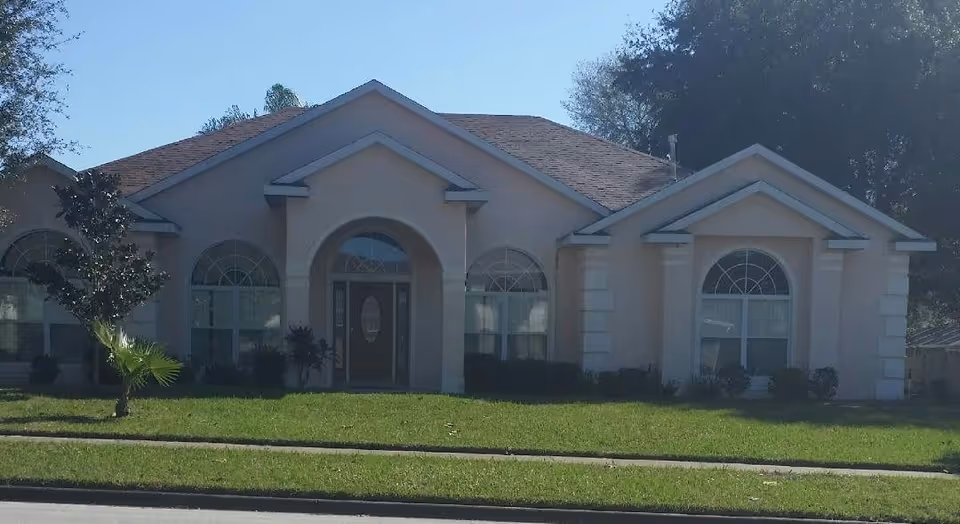 Front exterior view of a single-story residential building with a beige facade, arched windows, and a central entrance door with glass panels. The building is surrounded by a green lawn and a few small trees and bushes.