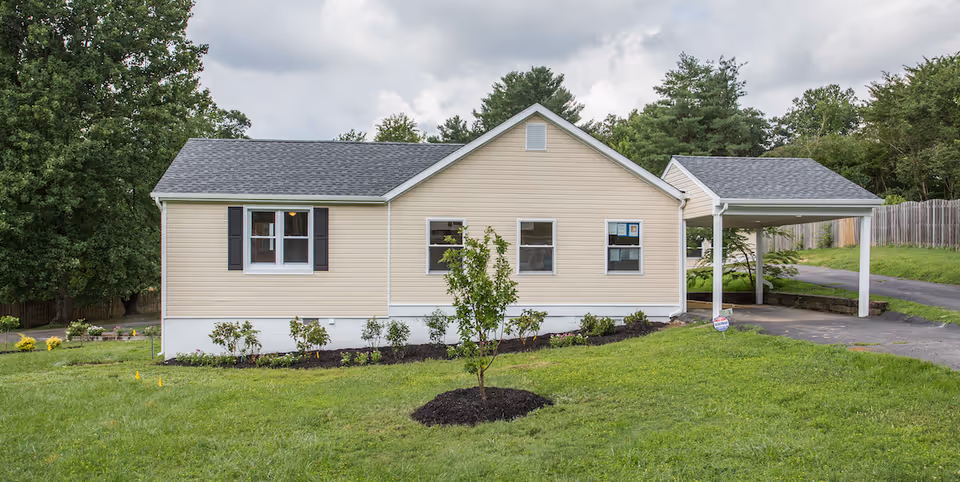 Single-story beige house with a carport, front lawn, planted shrubs and a young tree under a cloudy sky.